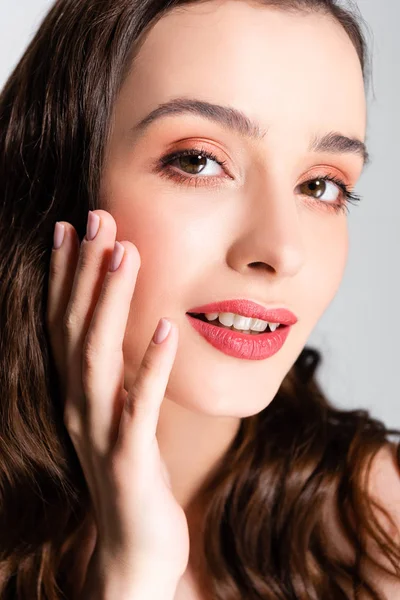 Hermosa mujer morena con rizos y maquillaje tocando la cara aislado en gris - foto de stock