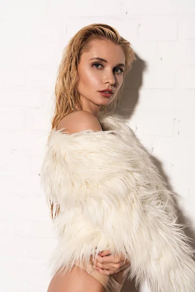 Sexy young woman with wet hair in white faux fur coat near white brick wall — Stock Photo