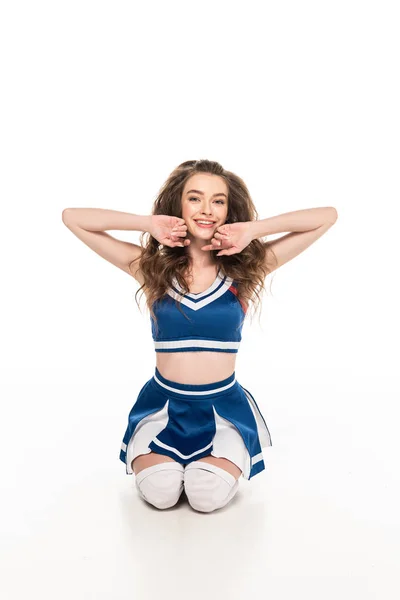 Sexy happy cheerleader girl in blue uniform sitting on floor and touching face isolated on white — Stock Photo