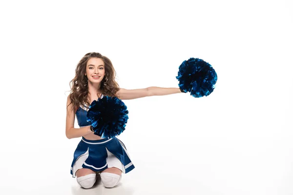 Menina cheerleader feliz em uniforme azul sentado com pompons no chão isolado no branco — Fotografia de Stock