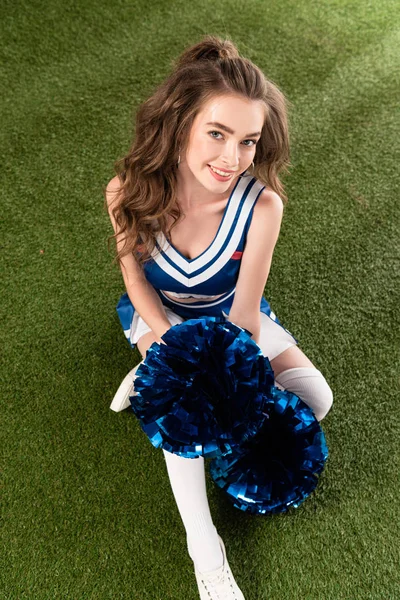 Vista aérea de chica bonita animadora en uniforme azul sentado con pompones en el campo verde - foto de stock