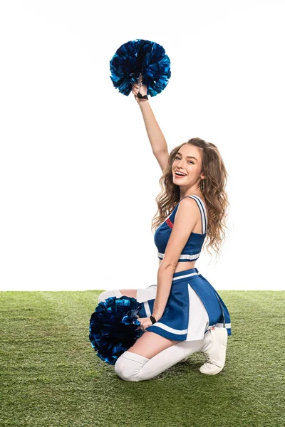 Smiling cheerleader girl in blue uniform sitting with pompoms on green field isolated on white — Stock Photo