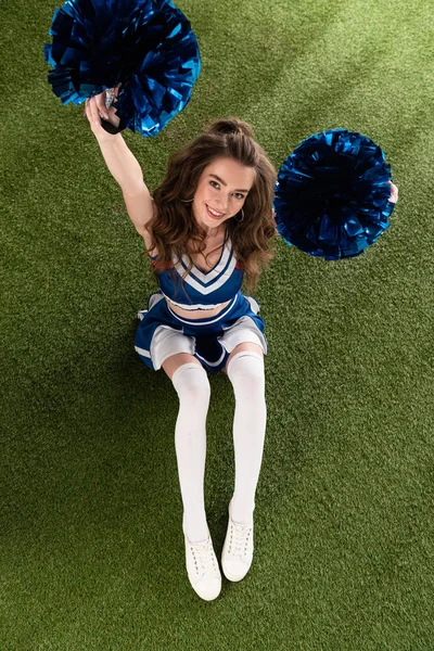 Vista dall'alto della ragazza sexy cheerleader in uniforme blu seduta con pompon sul campo verde — Foto stock