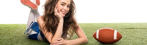 Smiling cheerleader girl in blue uniform lying near rugby ball on green field isolated on white, panoramic shot — Stock Photo