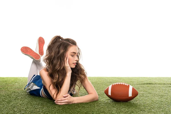 Cheerleader-Mädchen in blauer Uniform liegt neben Rugby-Ball auf grünem Feld isoliert auf weißem Grund — Stockfoto