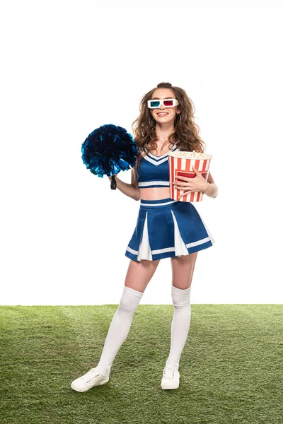 Fröhliches Cheerleader-Mädchen in blauer Uniform und 3D-Brille, stehend mit Popcorn und Bommel auf grünem Feld isoliert auf weißem Grund — Stockfoto