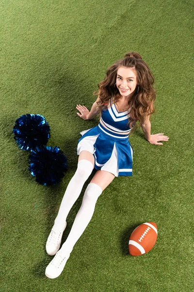 Vista aérea de chica animadora bonita en uniforme azul acostado con pompones y pelota de rugby en el campo verde - foto de stock