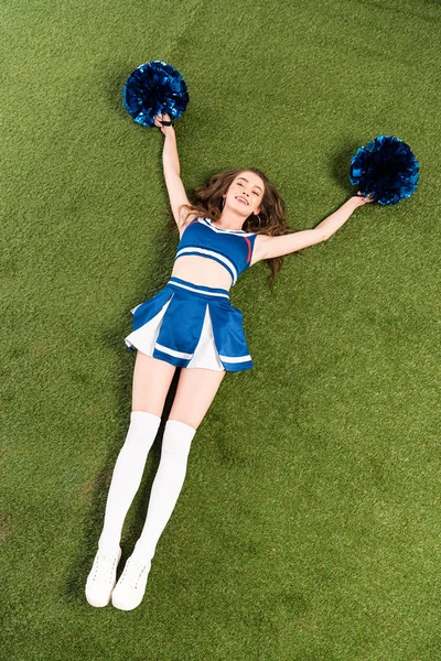 Overhead view of pretty cheerleader girl in blue uniform lying with pompoms on green field — Stock Photo
