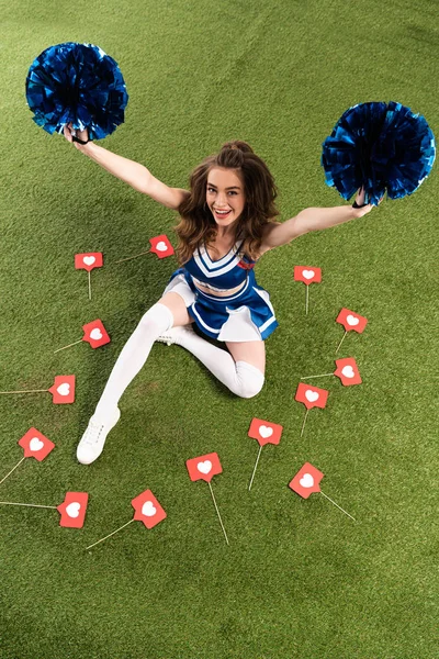 Overhead view of happy cheerleader girl in blue uniform sitting with pompoms on green field with scattered likes signs — Stock Photo