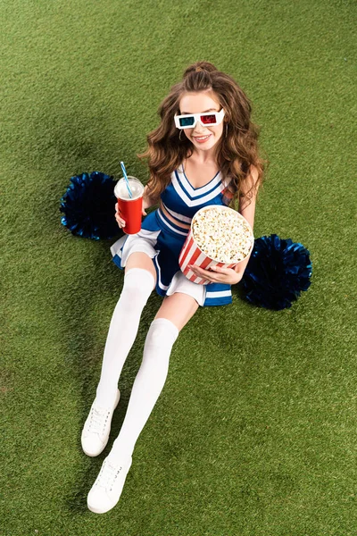 Overhead view of pretty cheerleader girl in blue uniform and 3d glasses with pompoms, popcorn and soda on green field — Stock Photo