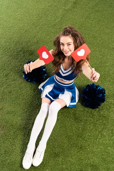 Vista aérea de chica animadora feliz en uniforme azul sentado con pompones y corazones de las redes sociales en el campo verde - foto de stock