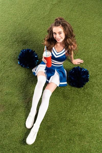 Vista aérea de menina bonita cheerleader em uniforme azul sentado com pompons e refrigerante no campo verde — Fotografia de Stock