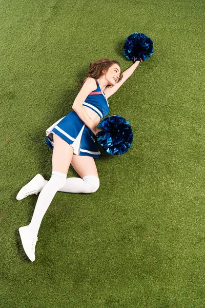 Overhead view of cheerleader girl in blue uniform lying with pompoms on green field — Stock Photo