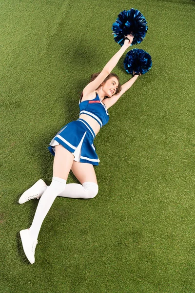 Vista aérea de animadora alegre chica en uniforme azul acostado con pompones en el campo verde - foto de stock