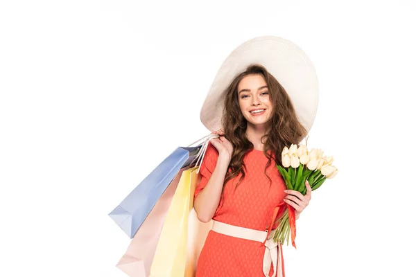 Sonriente mujer elegante en sombrero y vestido sosteniendo tulipanes blancos y bolsas aisladas en blanco - foto de stock