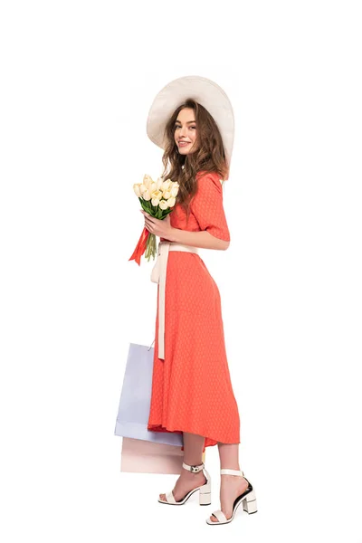 Glücklich elegante Frau in Hut und Kleid mit weißen Tulpen und Einkaufstaschen isoliert auf weiß — Stockfoto