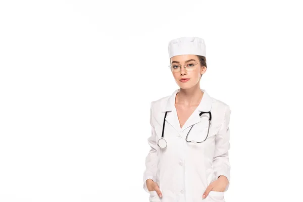Joven doctor en bata blanca con estetoscopio y manos en bolsillos aislados en blanco - foto de stock