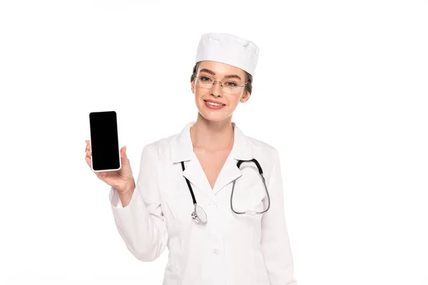 Joven médico feliz en bata blanca con estetoscopio mostrando teléfono inteligente con pantalla en blanco aislado en blanco - foto de stock