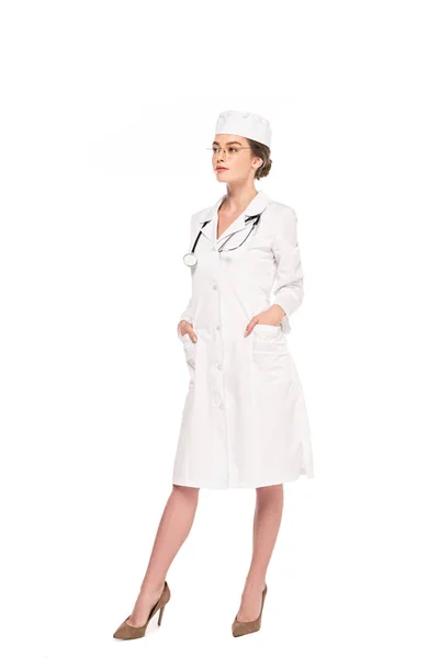 Piena vista del giovane medico in cappotto bianco con stetoscopio in posa con le mani in tasche isolate su bianco — Foto stock