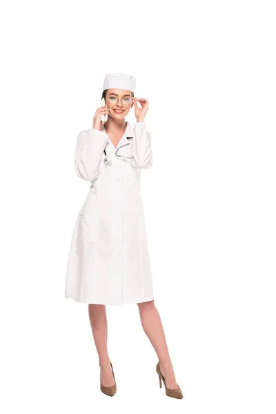 Full length view of young doctor in white coat with stethoscope talking on smartphone isolated on white — Stock Photo