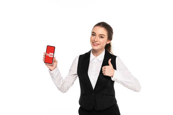 KYIV, UCRANIA - 7 DE ABRIL DE 2019: joven camarera feliz sosteniendo el teléfono inteligente con la aplicación de youtube y mostrando el pulgar hacia arriba aislado en blanco - foto de stock