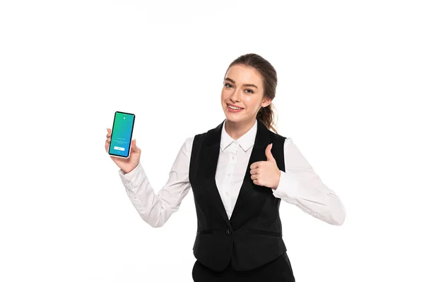 KYIV, UCRANIA - 7 DE ABRIL DE 2019: joven camarera feliz sosteniendo el teléfono inteligente con la aplicación de twitter y mostrando el pulgar hacia arriba aislado en blanco - foto de stock