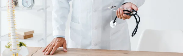 Plano panorámico del médico sosteniendo estetoscopio cerca de la mesa en la clínica — Stock Photo