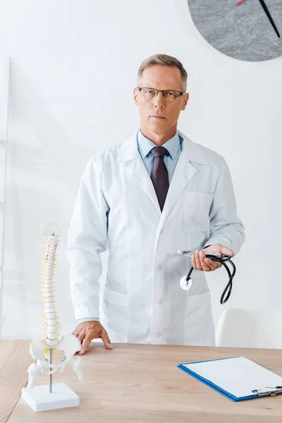 Handsome doctor in white coat standing and holding stethoscope near table — Stock Photo