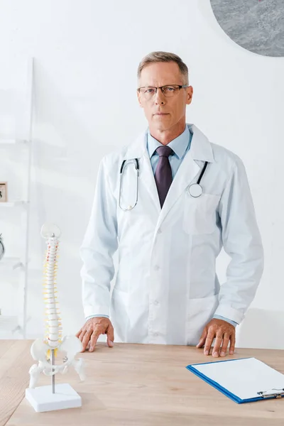 Médico bonito em óculos e casaco branco em pé perto da mesa — Fotografia de Stock