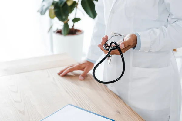 Vista cortada do médico segurando estetoscópio perto da mesa de madeira — Fotografia de Stock