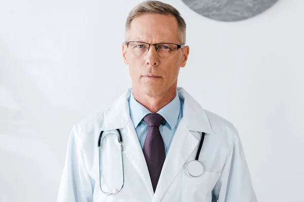 Gutaussehender Arzt mit Brille und weißem Mantel blickt in die Kamera — Stockfoto