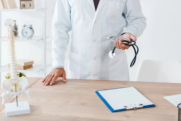 Vista recortada del médico sosteniendo estetoscopio cerca del portapapeles y la mesa - foto de stock