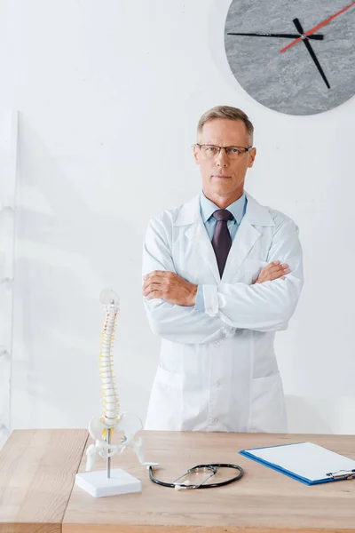 Médecin dans des lunettes debout avec les bras croisés près de la table — Photo de stock