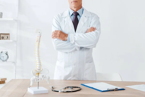 Vista cortada de médico de pé com braços cruzados perto de mesa de madeira — Fotografia de Stock