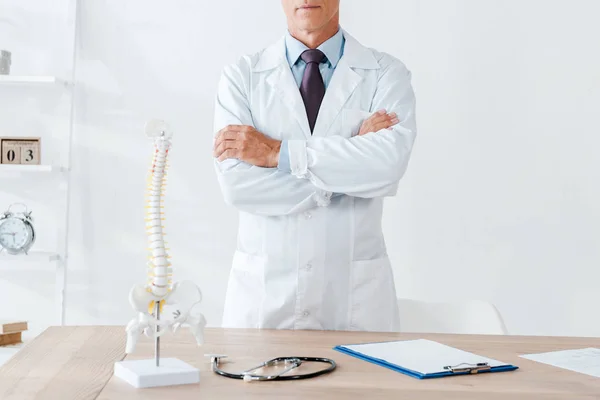 Vista recortada del médico de pie con los brazos cruzados cerca de la mesa de madera - foto de stock