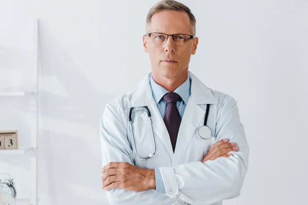 Médico con estetoscopio de pie con los brazos cruzados y mirando a la cámara - foto de stock