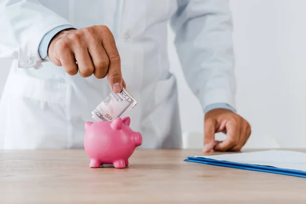 Обрезанный взгляд врача в белом халате положить доллар банкноты в копилку свиньи — стоковое фото