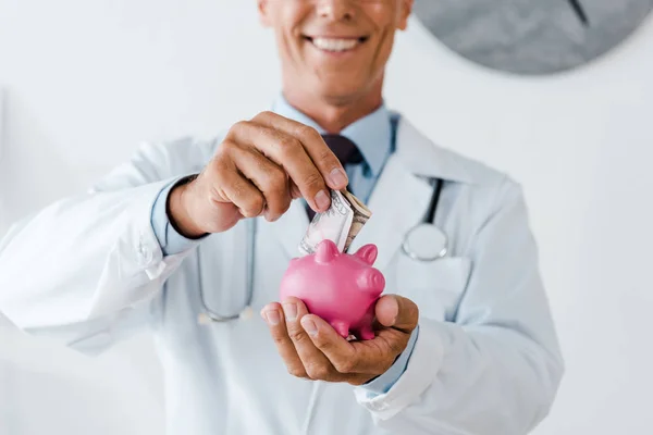 Abgeschnittene Ansicht eines fröhlichen Mannes in weißem Mantel, der Dollar-Banknoten in die Hand nimmt, während er Sparschwein hält — Stockfoto