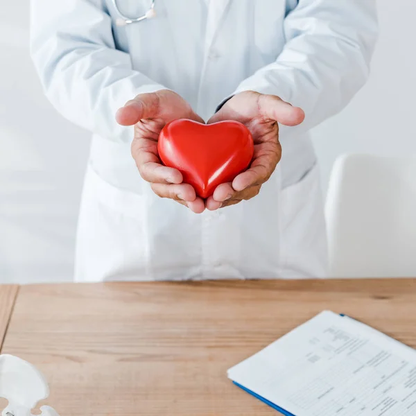 Vista recortada del médico sosteniendo modelo de corazón rojo cerca de la mesa - foto de stock