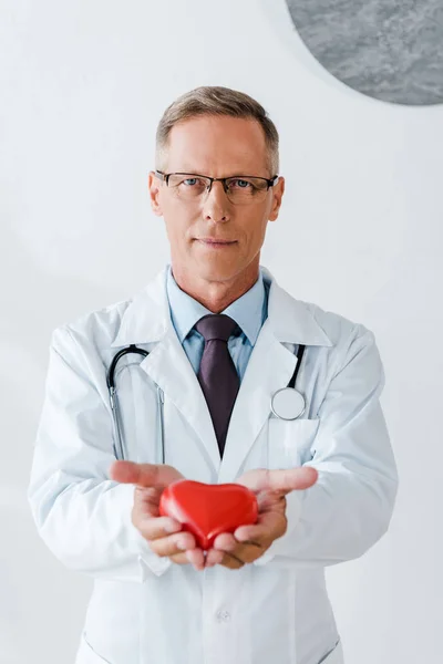 Guapo doctor en gafas y blanco abrigo sosteniendo rojo corazón modelo - foto de stock