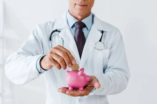 Ausgeschnittene Ansicht eines Arztes im weißen Kittel, der Münze in Sparschwein steckt — Stockfoto