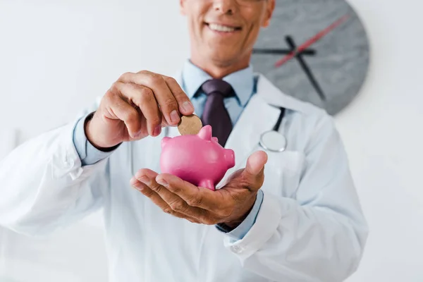 Ausgeschnittene Ansicht eines glücklichen Arztes im weißen Kittel, der Münze in Sparschwein steckt — Stockfoto