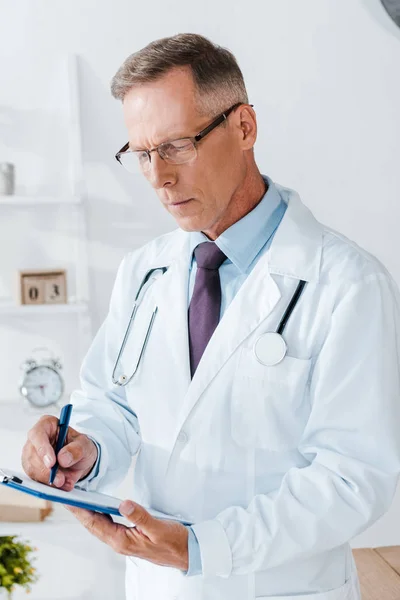 Guapo doctor en blanco abrigo y gafas escritura diagnóstico mientras sostiene portapapeles - foto de stock