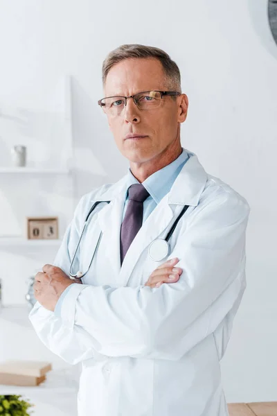 Médico serio en gafas y bata blanca de pie con los brazos cruzados - foto de stock