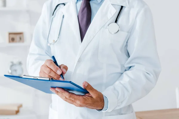 Vista recortada del médico en capa blanca sujetando portapapeles y diagnóstico de escritura - foto de stock