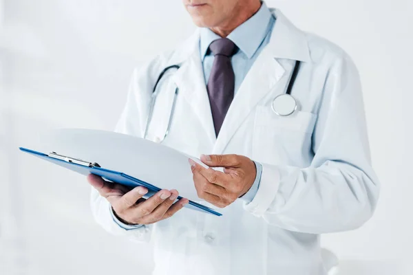 Vista cortada do médico em branco casaco segurando página e área de transferência na clínica — Fotografia de Stock