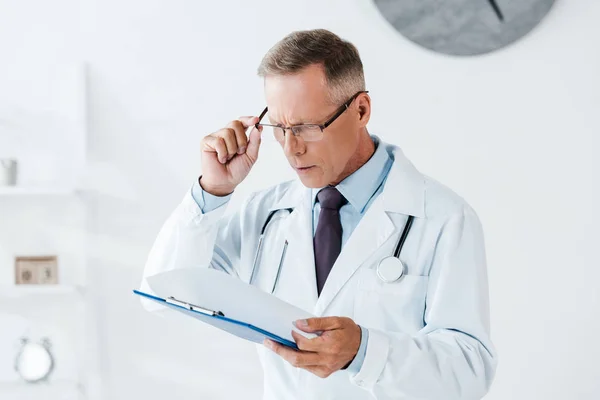 Schöner Mann im weißen Mantel, der in der Klinik beim Anblick des Klemmbretts eine Brille berührt — Stockfoto