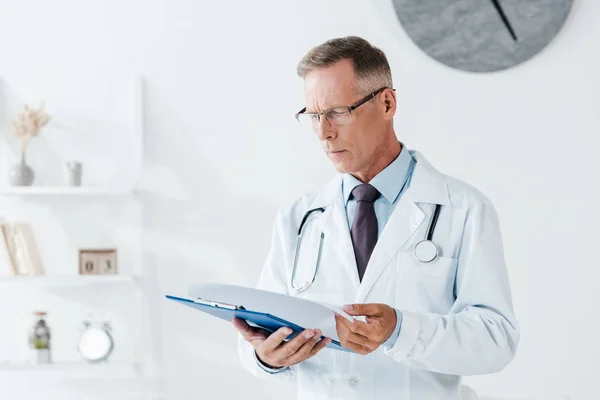 Schöner Mann im weißen Mantel schaut in Klinik auf Klemmbrett — Stockfoto