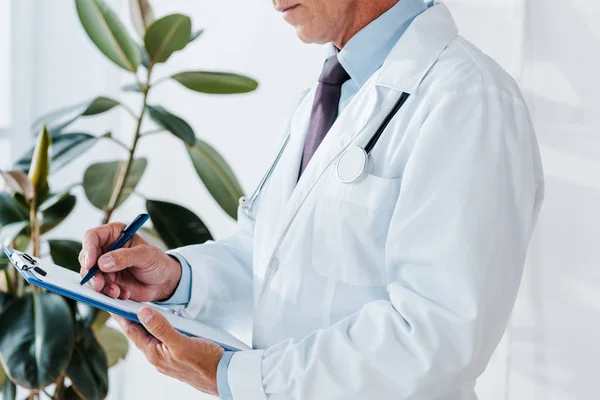 Cropped view of man in white coat writing diagnosis and holding clipboard — Stock Photo