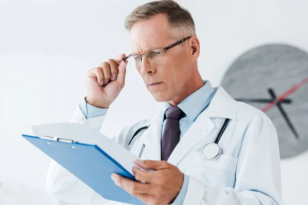 Selective focus of handsome doctor in white coat looking at clipboard and touching glasses in hospital — Stock Photo